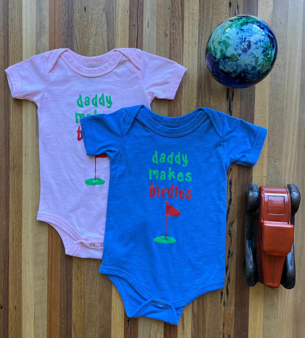 Putt Life Golf blue and pink baby onesies with the text 'Daddy makes birdies' and a green and red golf green and flag graphic on the front 1Zs
