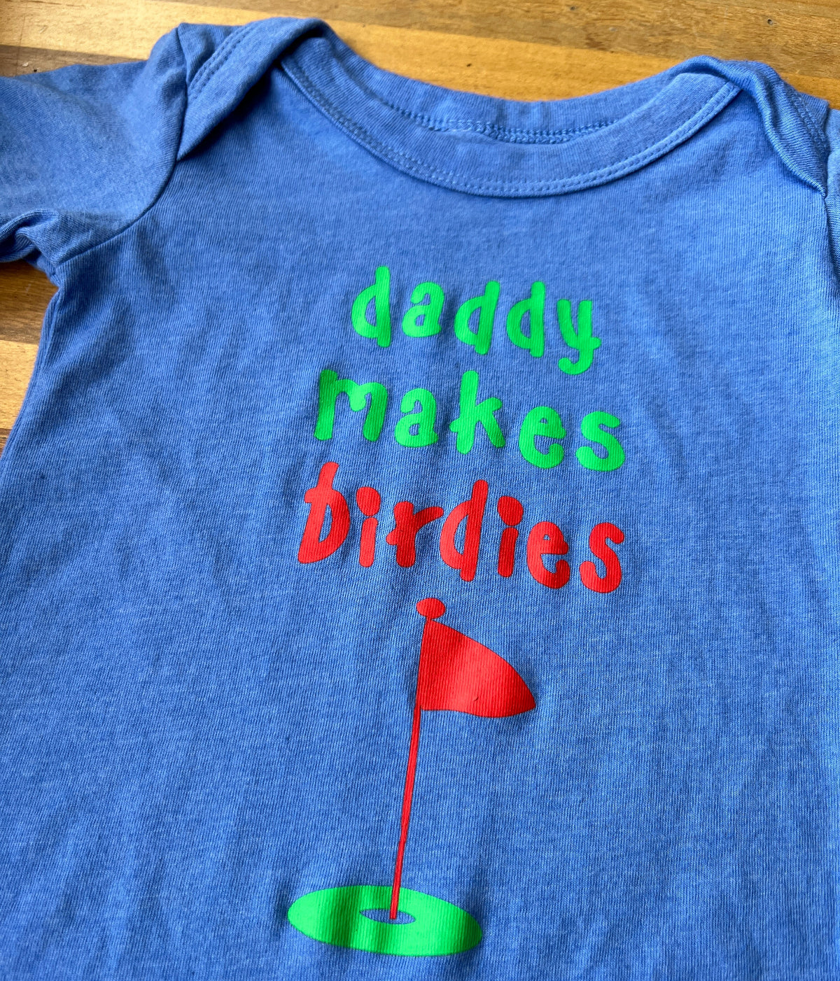 Putt Life Golf blue baby onesie with the text 'Daddy makes birdies' and a green and red golf green and flag graphic on the front 1Z close up
