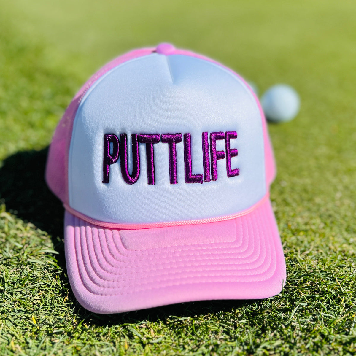 Putt Life Golf foam trucker hat—pink brim and pink mesh, white front with ‘PUTTLIFE’ in purple in the grass with golf ball in the background.