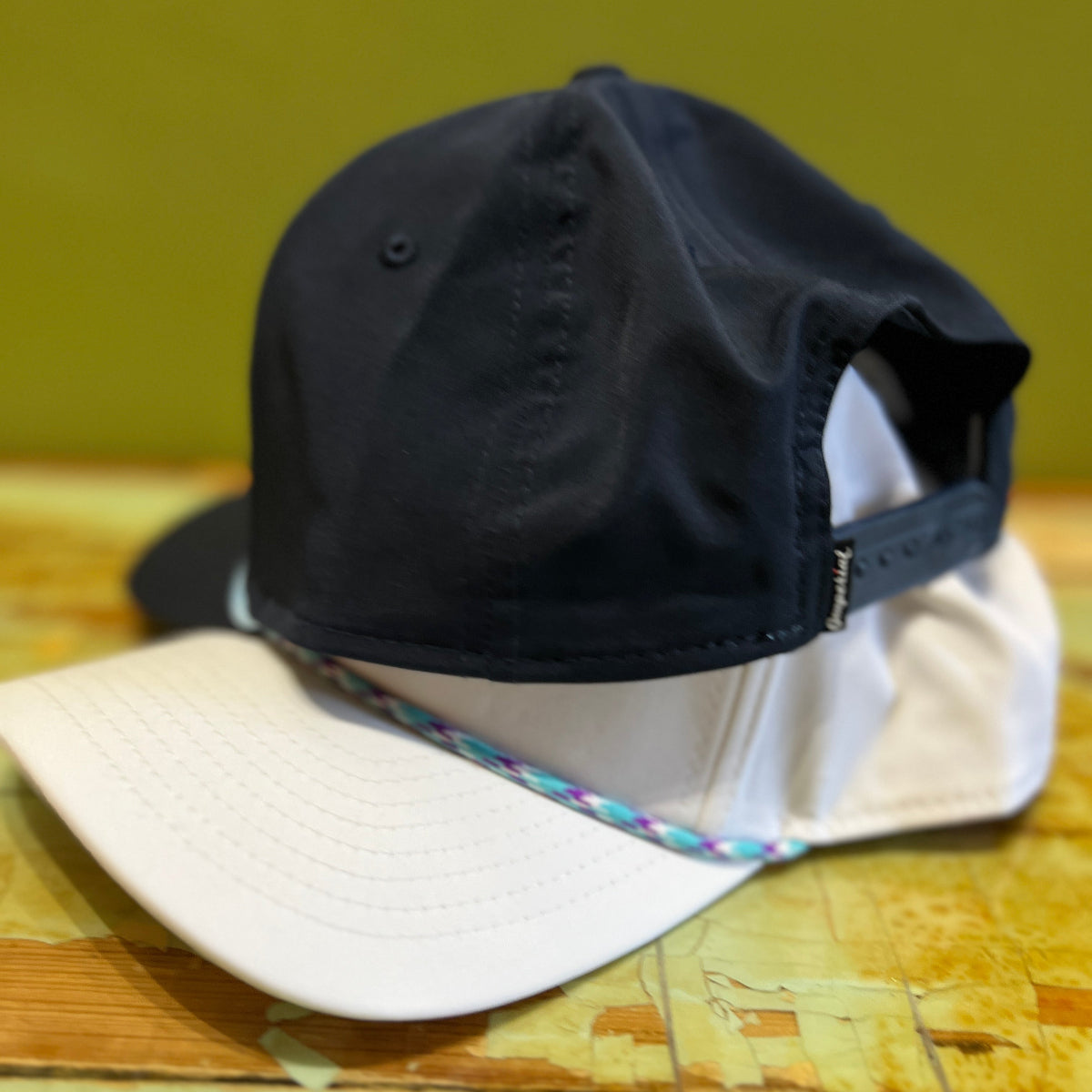 Putt Life Golf navy blue golf cap with logo and text "HAPPY PUTTING" with a light blue rope design around the brim, and white golf hat with light blue multi colored rope around brim show back of hat and snapback on a table