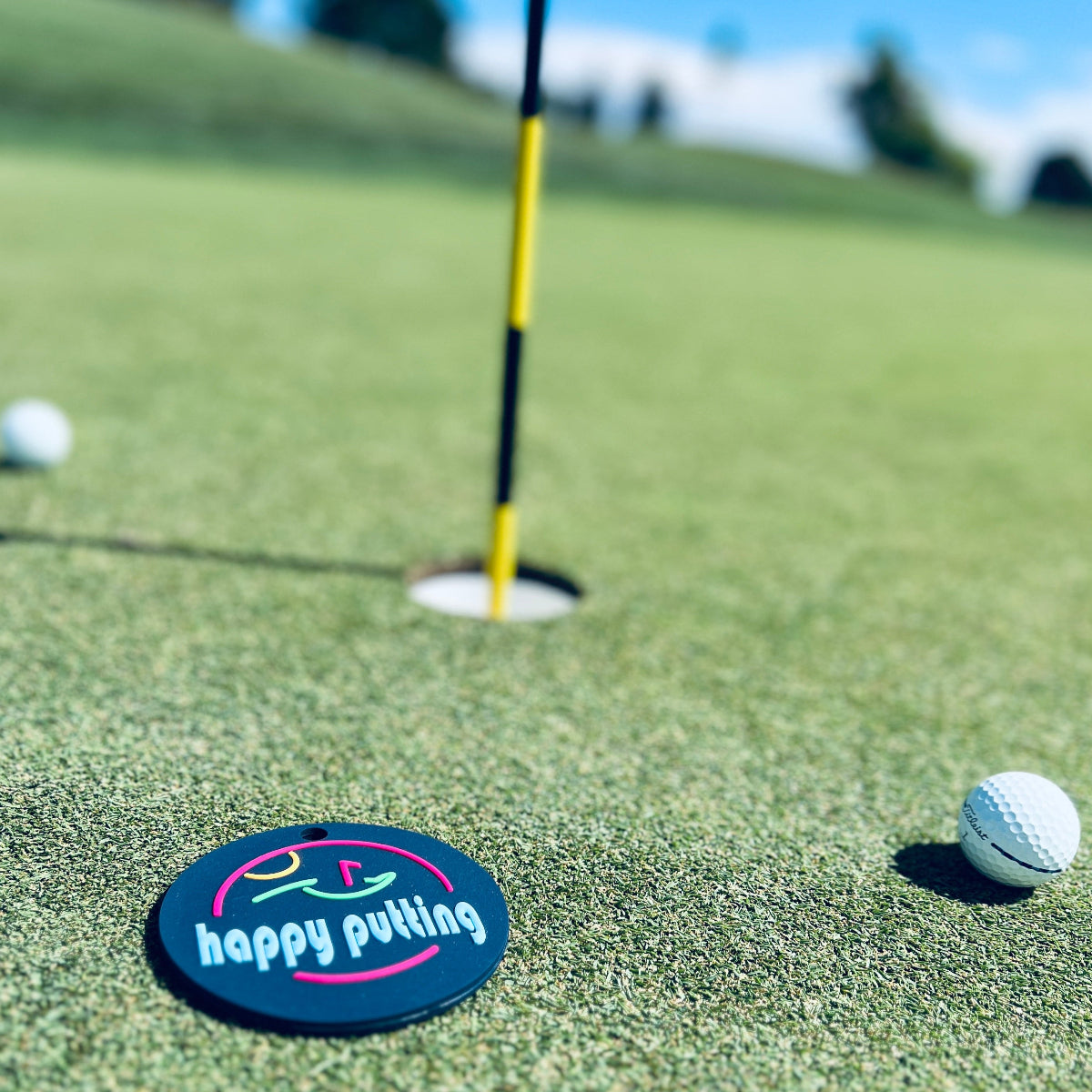 Putt Life Golf navy blue putting disc with the text "happy putting" in light blue and neon green putting green with neon pink flag and neon orange sun, lying on a green golf course putting green next to a golf hole and yellow flag stick with white golf balls in the background