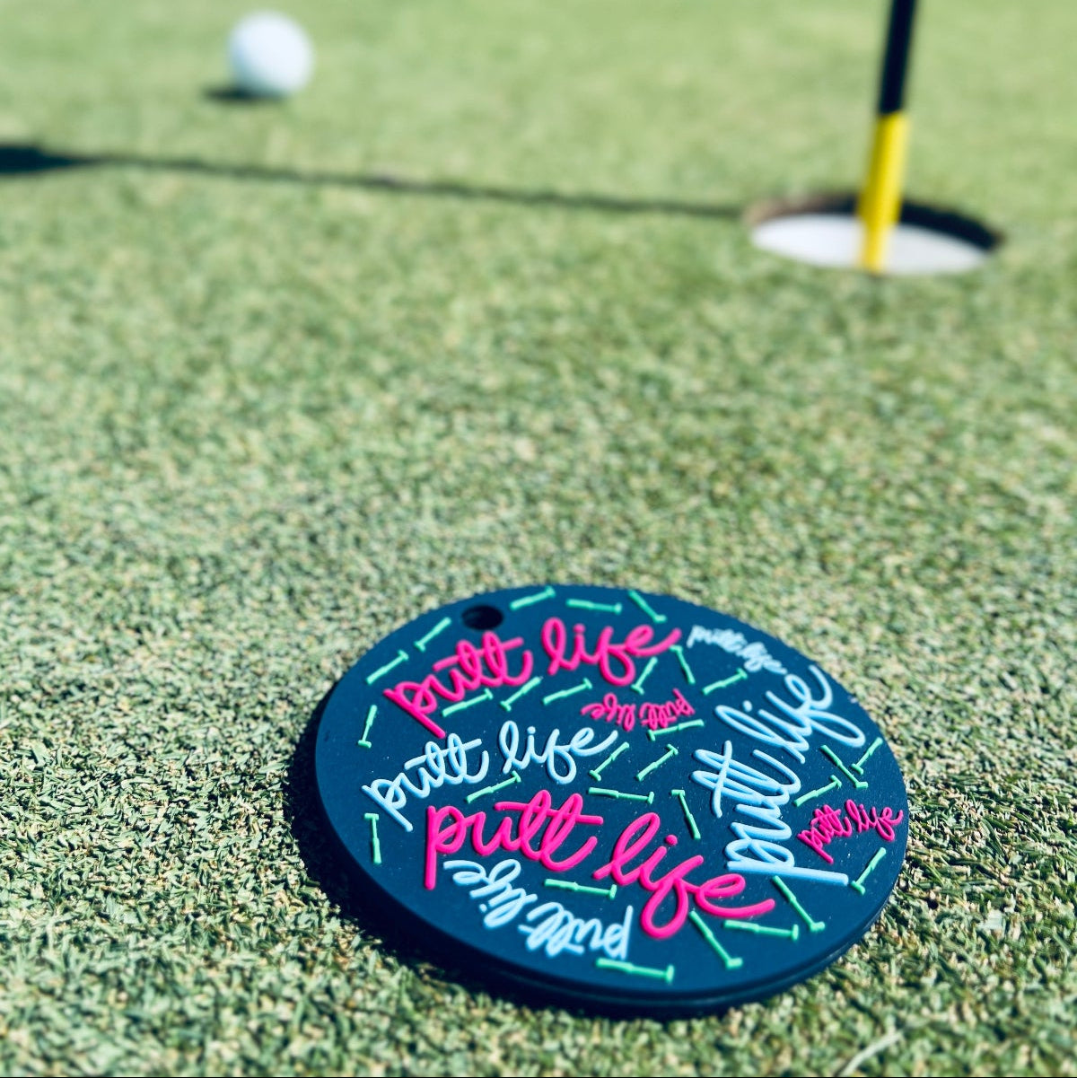 Putt Life Golf navy blue putting disc with pink and light blue putt life logos and neon green putters on it, lying on a green golf course putting green next to a golf hole and yellow flag stick with white golf ball in the background