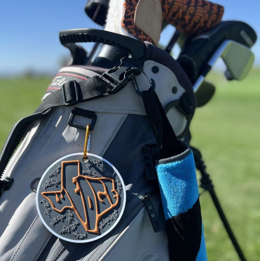Golf club bag with a decorative tag putting disc featuring a texas wedge putt life golf logo on a golf bag in a grassy field